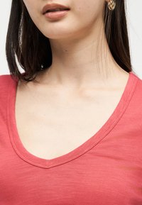 Γυναίκα με ίσια σκούρα μαλλιά που φοράει κοραλλί μπλούζα με V στο λαιμό και χρυσα σκουλαρίκια σε σχήμα στριφτών κρίκων, εμφανίζεται από το πηγούνι μέχρι το πάνω μέρος του στήθους.