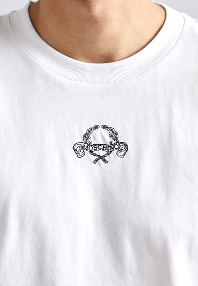 Balta medvilnė marškinėliai su apvaliame kakle, su juodu siuvinėjimu logotipo ir dekoratyviais elementais viduryje. Lygi tekstūra.