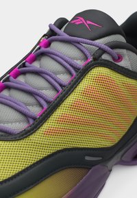 Γκρι και μαύρο αθλητικό παπούτσι με μωβ κορδόνια, κίτρινες πλευρικές επιφάνειες από δίχτυ, ροζ λογότυπο στη γλώσσα και λεπτομέρειες στη σόλα σε μωβ και μαύρο χρώμα.