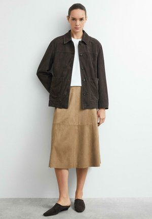 Frau steht vor einer weißen Wand, trägt eine dunkelbraune, geknöpfte Jacke, ein weißes Top, einen hellbraunen Midirock und dunkle Slipper.
