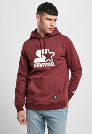 Bordeauxfarbener Hoodie mit einem weißen Logo, ausgestattet mit einer Vordertasche, einer Kapuze mit Kordelzug und gerippten Bündchen. Hergestellt aus weichem, strukturiertem Stoff.