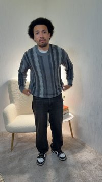 Homme avec une coiffure afro ajustant un pull gris rayé dans une pièce moquettée avec un fauteuil blanc et une table d'appoint avec des livres.