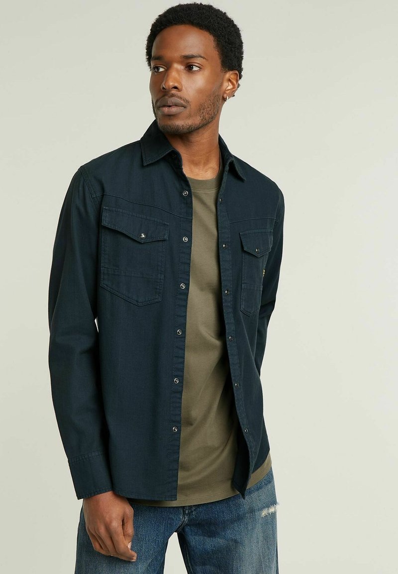 Camicia button-up blu scuro con due tasche sul petto, design semplice e maniche lunghe, indossata sopra una maglietta di colore verde oliva chiaro.