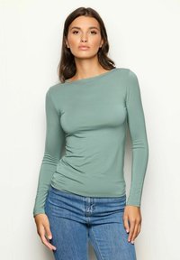 Langärmliges Oberteil in gedämpftem Grün, aus weichem Stoff gefertigt. Enganliegendes Design mit rundem Halsausschnitt, kombiniert mit Jeans. Glatte Textur.