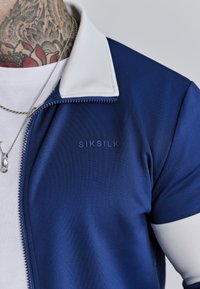 Mornarsko plavi zip-up kaput s bijelim ovratnikom, napravljen od glatkog materijala. Ima logo branda u mornarskoj boji na prsima i vidljivi detalj sa zatvaračem.