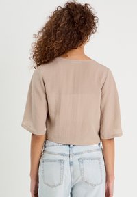 Frau mit lockigem Haar trägt ein beiges, locker sitzendes Kurzarmshirt und hellblaue, hoch taillierte Jeans, blickt von hinten vor einem schlichten Hintergrund.