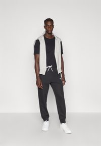 Γκρι αμάνικο φούτερ με κουκούλα πάνω από μαύρο T-shirt, συνδυασμένο με μαύρη φόρμα και λευκά αθλητικά παπούτσια. Σημαντική λεπτομέρεια λογότυπου στο παντελόνι.