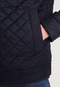Navy gewatteerde jas met een ruitpatroon, voorzien van een zijzak en ribgebreide accenten op de manchetten en de zoom. Soepele, lichte stof.