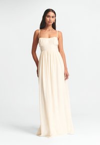 Vestido largo de color crema hecho de una tela ligera. Presenta un escote cuadrado, tirantes delgados, un cuerpo fruncido y una falda fluida y amplia.