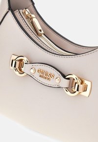Borsa beige con materiale testurizzato, dettagli in metallo dorato e un'etichetta in pelle con il marchio. Caratterizzata da un compartimento con zip sulla parte superiore.
