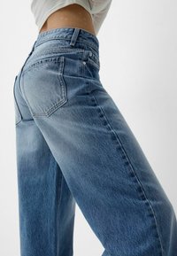Jeans din denim albastru deschis, cu o croială lejeră, având un design cu cinci buzunare, ușoară decolorare și cusături vizibile pe margini.