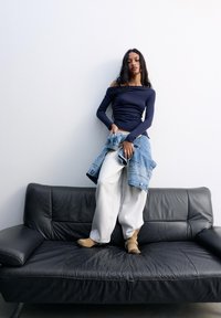 Marinblå långärmad topp, ljusgrå sweatpants, beige ankelstövlar och jeansjacka på en svart lädersoffa mot en vit vägg.