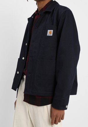 Man draagt een donkerblauw Carhartt-jack over een donkerrood geruit overhemd en gebroken-witte broek, staand tegen een effen witte achtergrond.