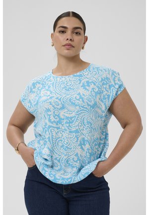 Femeie care poartă un top cu mâneci scurte, cu model paisley albastru și alb, și blugi închiși la culoare, stând cu mâinile în buzunare, pe un fundal simplu.