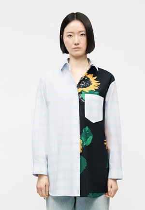 Woman with straight black hair wears a long-sleeve shirt split vertically: light blue check on one side, sunflower print on black on the other.