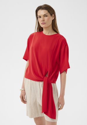 Žena má na sebe červený krátky rukáv s viazaním vpredu a béžové kraťasy po kolená, stojaca na bielom pozadí.