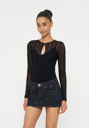 Jeune femme avec les cheveux en chignon portant un haut en dentelle noire à manches longues avec une encolure en trou de serrure et une jupe courte en jean foncé.