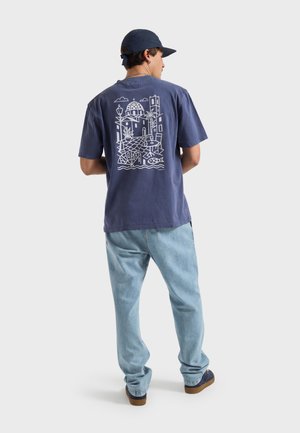 Hombre con gorra azul marino, camiseta azul marino con gráfico blanco de paisaje urbano en la espalda, vaqueros azul claro y zapatos oscuros, de pie de espaldas.