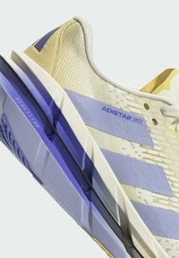 Scarpe da corsa beige chiaro realizzate in rete e materiali sintetici, con accenti blu-viola, suola testurizzata e tre strisce audaci.