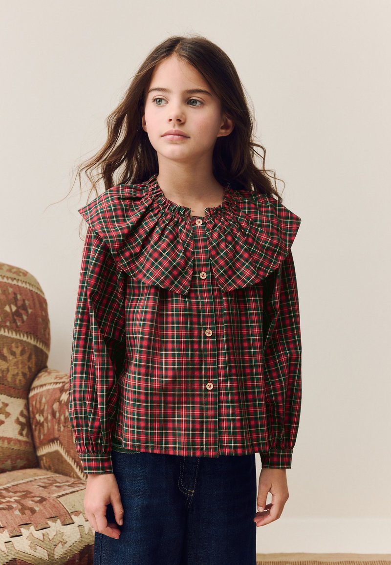 Chemise à carreaux rouges et verts avec boutons, grand col à volants et manches élastiquées, associée à un pantalon en denim foncé.