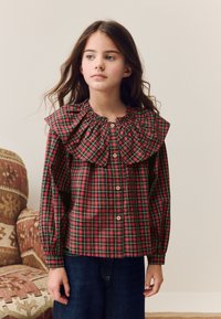 Chemise à carreaux rouges et verts avec boutons, grand col à volants et manches élastiquées, associée à un pantalon en denim foncé.