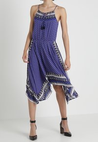 Bezramenné modré šaty s geometrickými a květinovými vzory, asymetrickým lemem a šňůrkou s třásněmi u výstřihu, kombinované s černými sandály na kotníku.