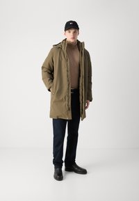 Olivgrüne gepolsterte Parkajacke, Frontreißverschluss, normale Passform. Brauner Rollkragenpullover darunter, marineblaue Hose und schwarze Lederstiefel. Schwarze Mütze.