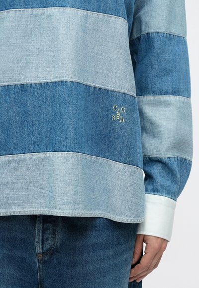 Chemise en denim à rayures horizontales bleu clair et bleu foncé, avec un logo "CLOSED" brodé en jaune en bas à droite. Manches longues avec poignets blancs.