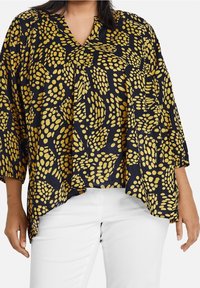 Czarna bluzka w żółte kropki, dekolt w serek, rękawy o długości trzech kwart, luźny krój i fakturowany materiał. W zestawie z białymi spodniami.