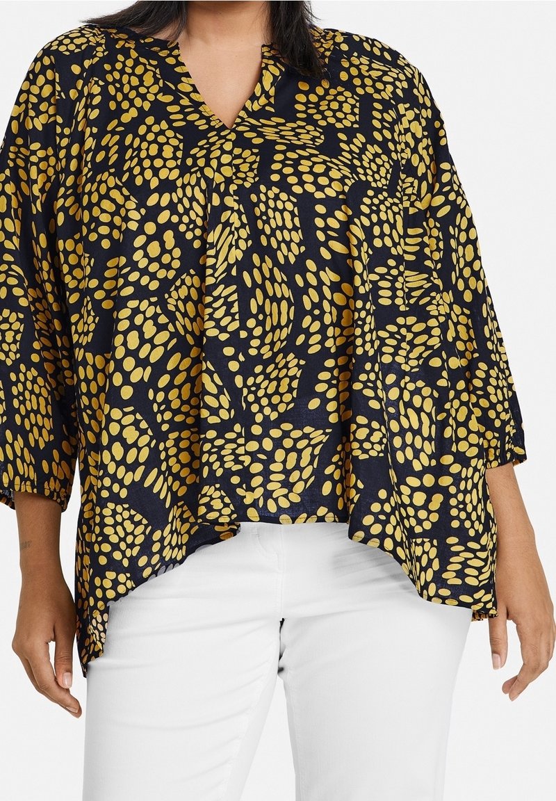 Czarna bluzka w żółte kropki, dekolt w serek, rękawy o długości trzech kwart, luźny krój i fakturowany materiał. W zestawie z białymi spodniami.