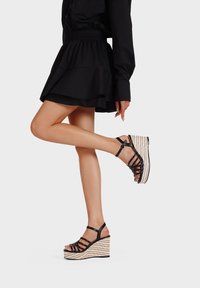 Sandali con zeppa neri a strappo con una piattaforma intrecciata elevata e cinghia alla caviglia regolabile. Texture liscia con motivo a strisce orizzontali.