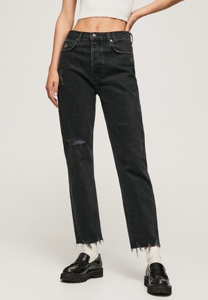 Vrouw draagt zwarte, hoog getailleerde versleten spijkerbroek met rafelige zoom, witte sokken, zwarte loafers en een cropped wit topje, staand tegen een beige achtergrond.