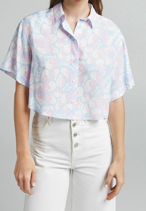 Femme portant une chemise courte à boutons rose pastel et bleue avec des motifs yin-yang et du texte, associée à un jean blanc taille haute.