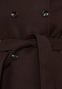 Manteau en laine marron foncé avec des boutons noirs et une ceinture en tissu nouée à la taille.