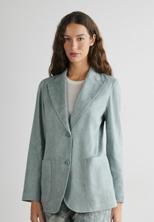 Kvinde med bølget brunt hår iført en lyseblå blazer over en hvid top, stående mod en ensfarvet hvid baggrund.