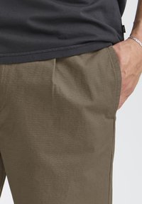 Pantalon kaki texturé avec un motif subtil en quadrillage, doté d'une poche latérale et d'une coupe décontractée, porté avec un t-shirt gris foncé.