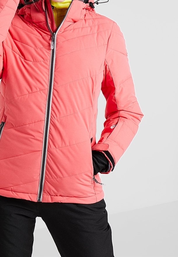 Personne portant une veste matelassée rose corail avec fermeture éclair devant et des gants noirs, main dans une poche zippée, pantalon noir, fond clair uni.