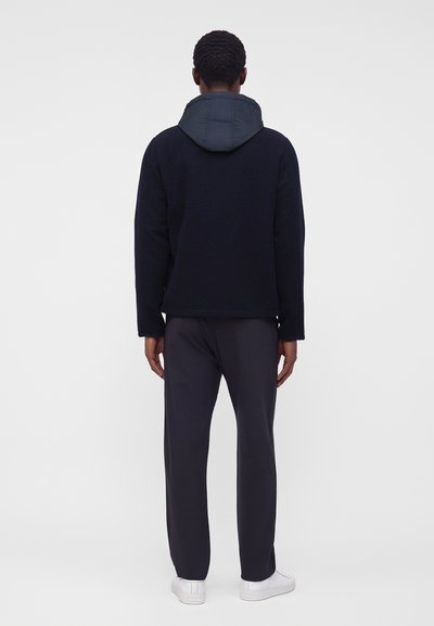 Personne debout, vue de dos, portant une veste à capuche noire, un pantalon foncé et des baskets blanches sur un fond clair uni.