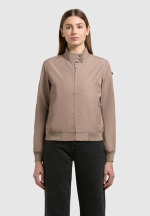 Femeie purtând o jachetă bomber cu fermoar, de culoare maro deschis, cu manșete și tiv striate, combinată cu jeansi negri cu talie înaltă, stând în picioare, cu fața către înainte.