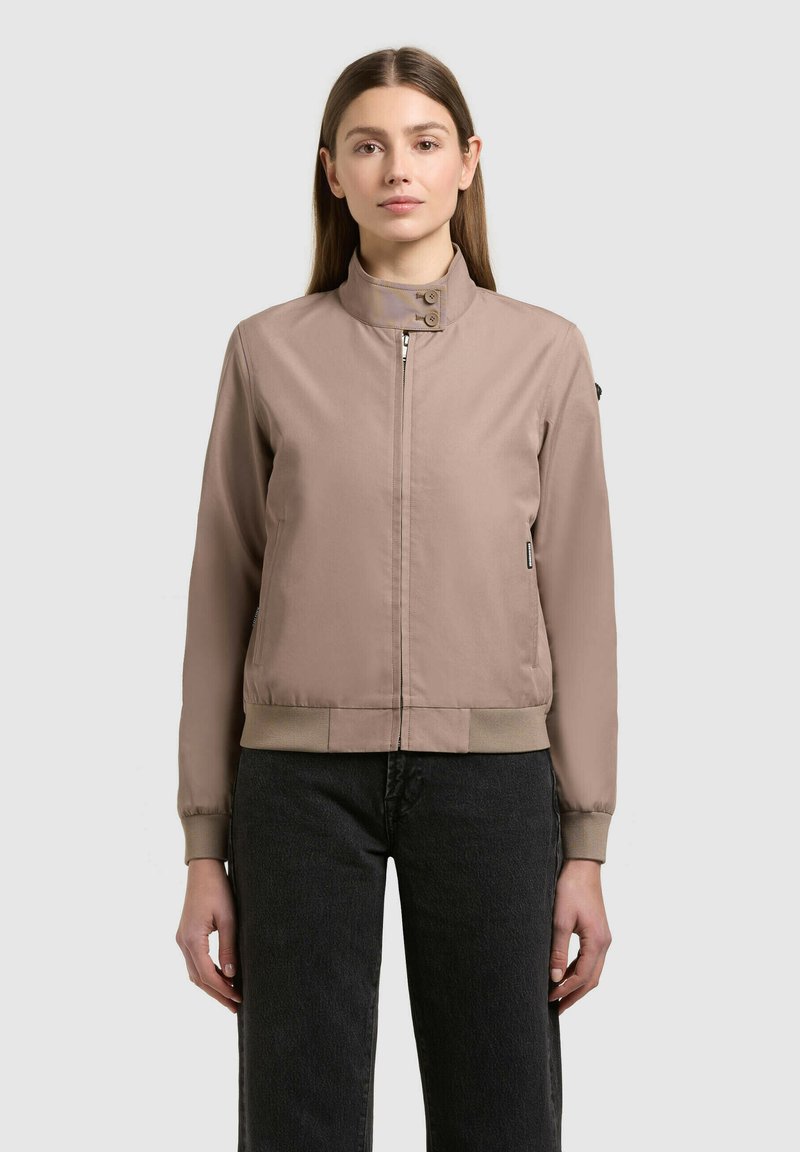 Femme portant une veste bomber marron clair à fermeture éclair avec des poignets et un ourlet côtelés, associée à un jean noir taille haute, debout face à l'avant.