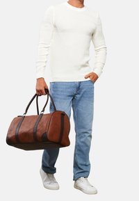 Braune Lederreisetasche mit dunklen Akzenten, runder Form, zwei Griffen und seitlichen Streifen. Leicht strukturierte Oberfläche, getragen von einer Person in Jeans und Pullover.