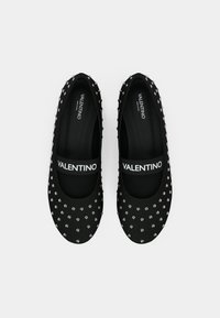 Sapatilhas pretas com um tecido texturizado apresentando tachas prateadas. Uma alça preta com a marca "VALENTINO" atravessa a parte superior. Design de bico arredondado.