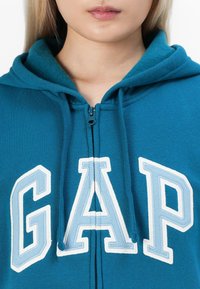 Teal zip-up hoodie featuring a kangaroo pocket, drawstring hood, and "GAP" logo in white and light blue embroidery. Soft texture.