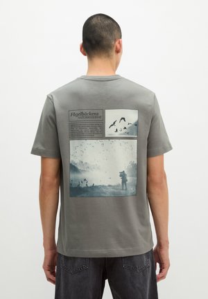 Mann trägt graues T-Shirt mit schwarz-weißen Naturfotos von Vögeln und einer Person mit Fernglas auf dem Rücken.