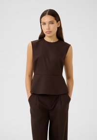 Smeđi bezrukavni peplum top od glatkog materijala, s oblikovanim gornjim dijelom i lepršavim donjim dijelom. Uparen s odgovarajućim smešnim hlačama.