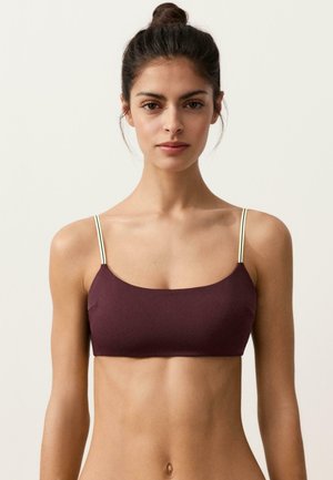 Młoda kobieta z włosami związanymi w kok, ubrana w bordowy krótki top z cienkimi kontrastującymi ramiączkami, stojąca na jasnym, jednolitym tle.