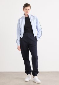 Svetlomodrá bunda na zips s čiernymi akcentmi, skombinovaná s čiernym topom a námorníckymi teplákmi. Biely tenisky dopĺňajú outfit. Hladká textúra po celom oblečení.