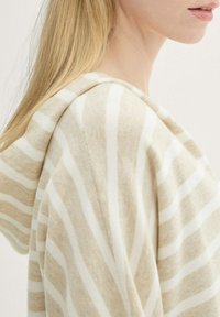 Pull à capuche rayé beige et blanc, avec une texture douce et un design drapé. La capuche présente une finition nette avec des coutures minimales.