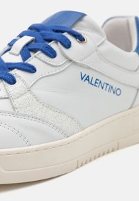White leather sneaker with blue laces and accents. Features a cracked texture on the side and embossed logo branding. Rubber outsole.