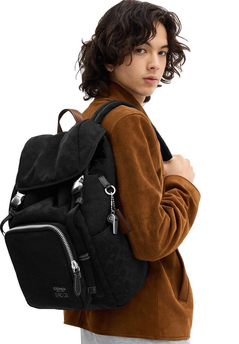 Young person with curly hair wearing a brown jacket and carrying a black Coach backpack over one shoulder.